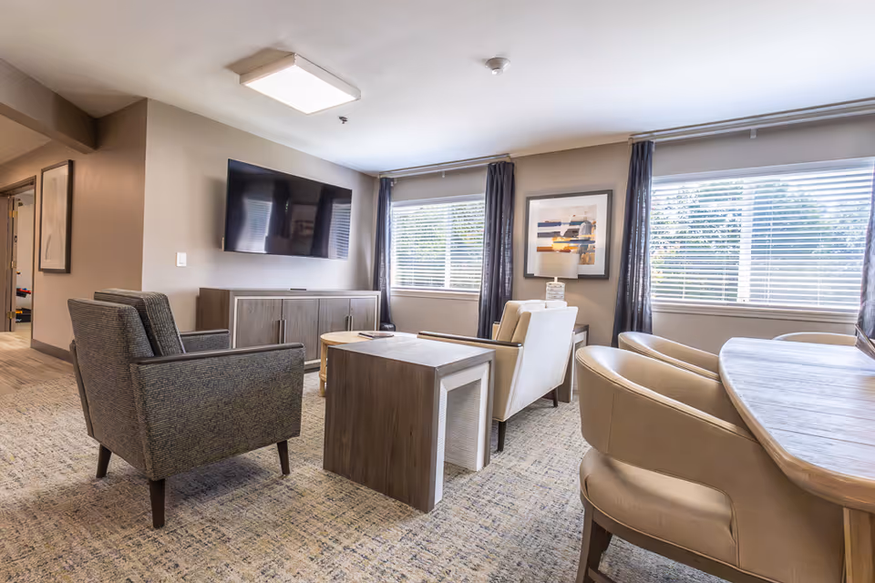 Furnished communal living room with upholstered chairs, a wall-mounted TV, coffee table, and large windows with blinds.