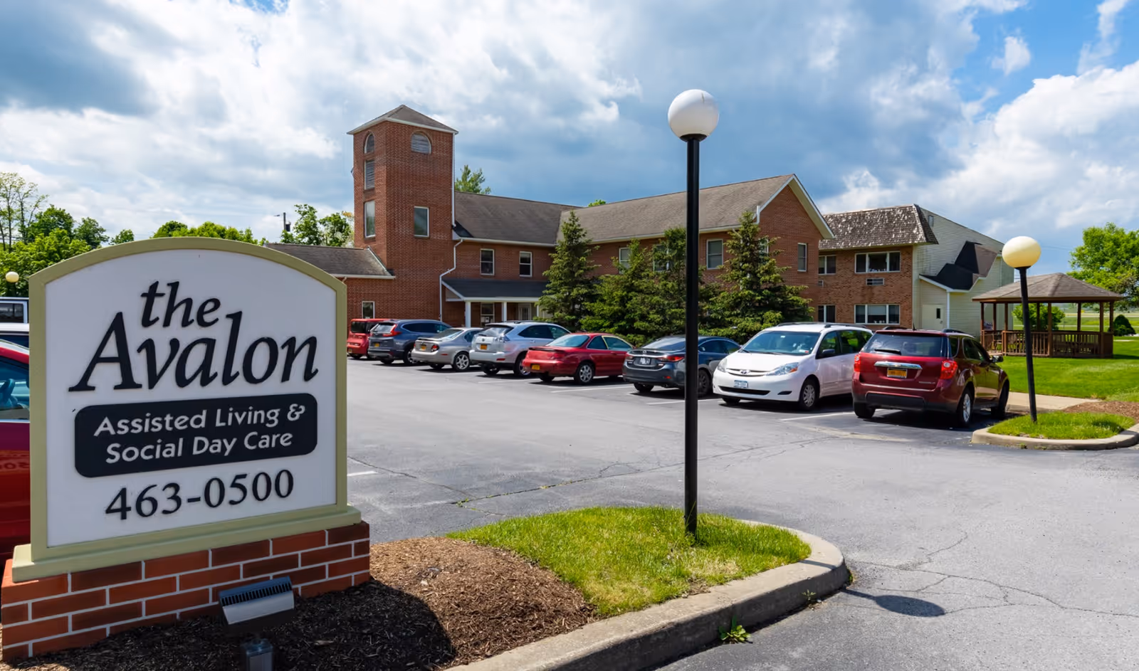 Exterior view of Avalon Assisted Living facility showing a brick building with multiple windows, a parking lot with several parked cars, a grassy area with lamp posts, and a gazebo in the background under a partly cloudy sky. A large sign in the foreground reads 'the Avalon Assisted Living & Social Day Care 463-0500'.
