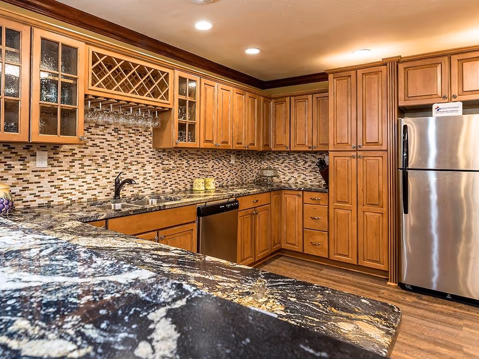 A modern kitchen with wooden cabinets, a mosaic tile backsplash, granite countertops, a stainless steel refrigerator, a dishwasher, and a double sink. There are wine glass holders with glasses hanging under the upper cabinets and recessed ceiling lights illuminating the space.