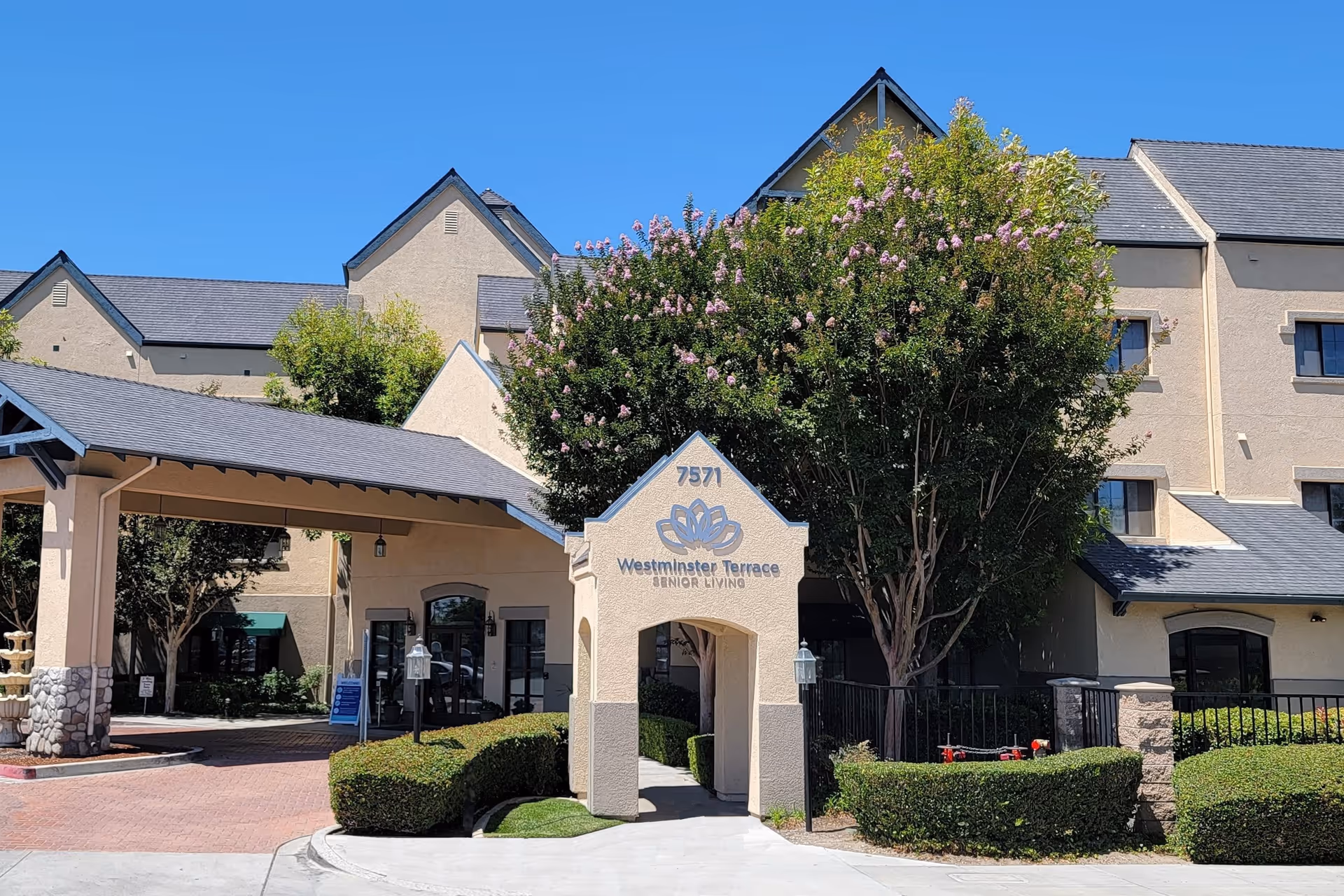Front entrance and facade of Westminster Terrace senior living with an arched entry sign, covered drive-through, and landscaped shrubs and trees.