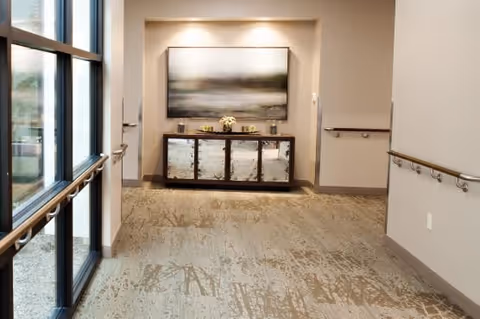 Carpeted interior corridor with handrails, large windows on the left, and a console table topped with artwork at the far end.