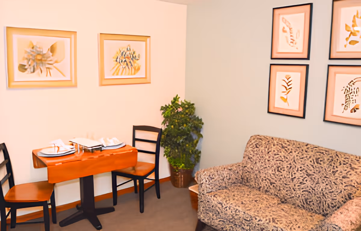 Small living area with a patterned sofa, a two-seat dining table set with plates, framed botanical artwork on the walls, and a potted plant.