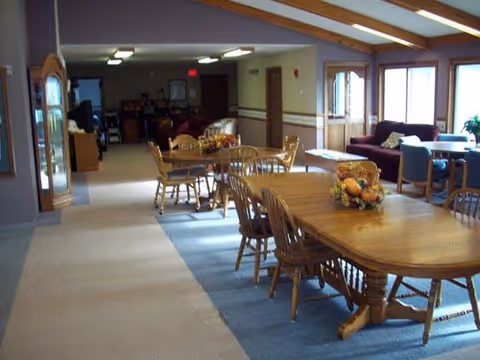 Communal dining area with wooden tables and chairs, floral centerpieces, and a seating area with couches near windows.