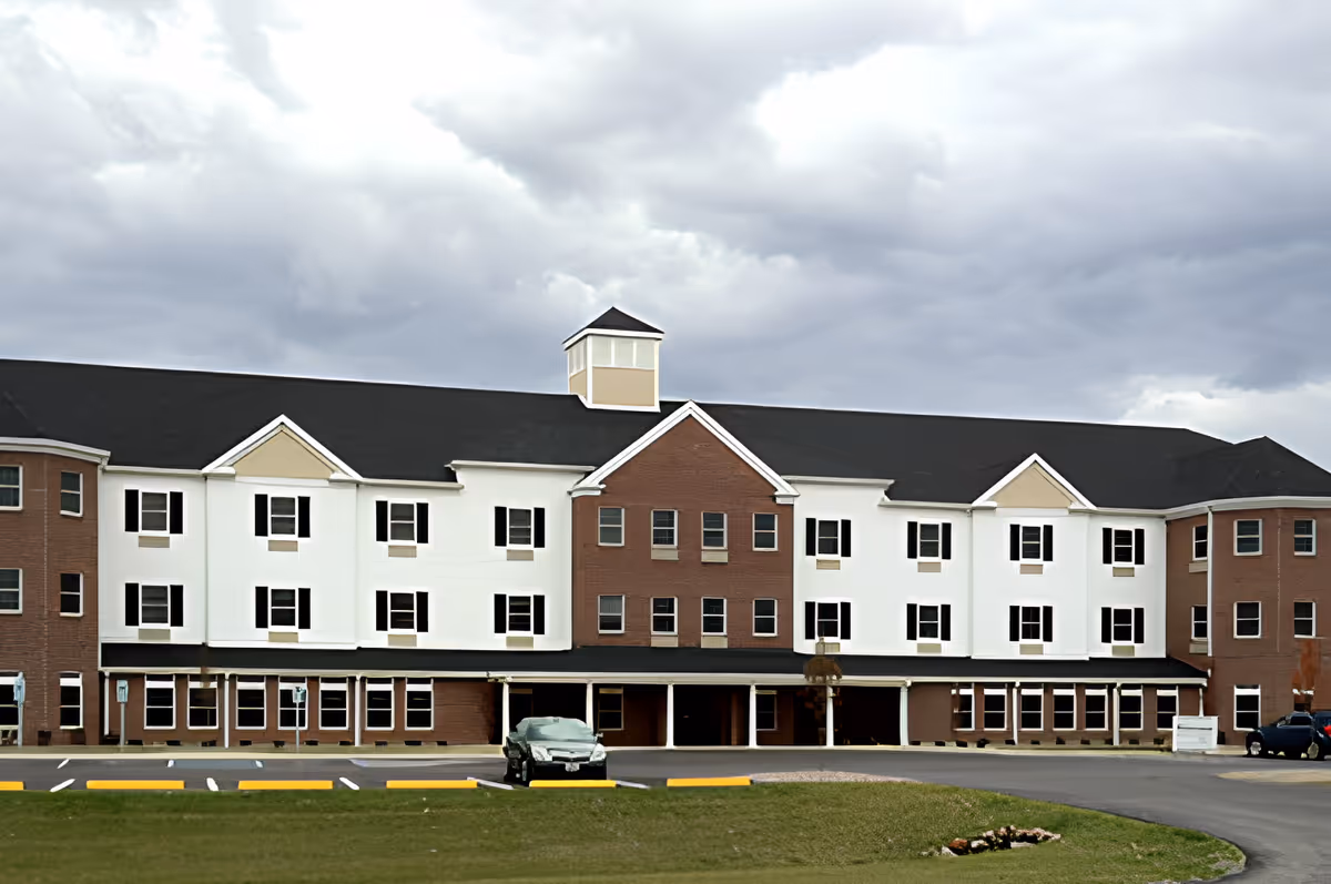 Front exterior view of a large, multi-story assisted living facility building with a combination of brick and white siding, black shutters on the windows, and a dark roof under a cloudy sky. There is a parking lot with a few cars in front of the building and a small grassy area with a flower bed.