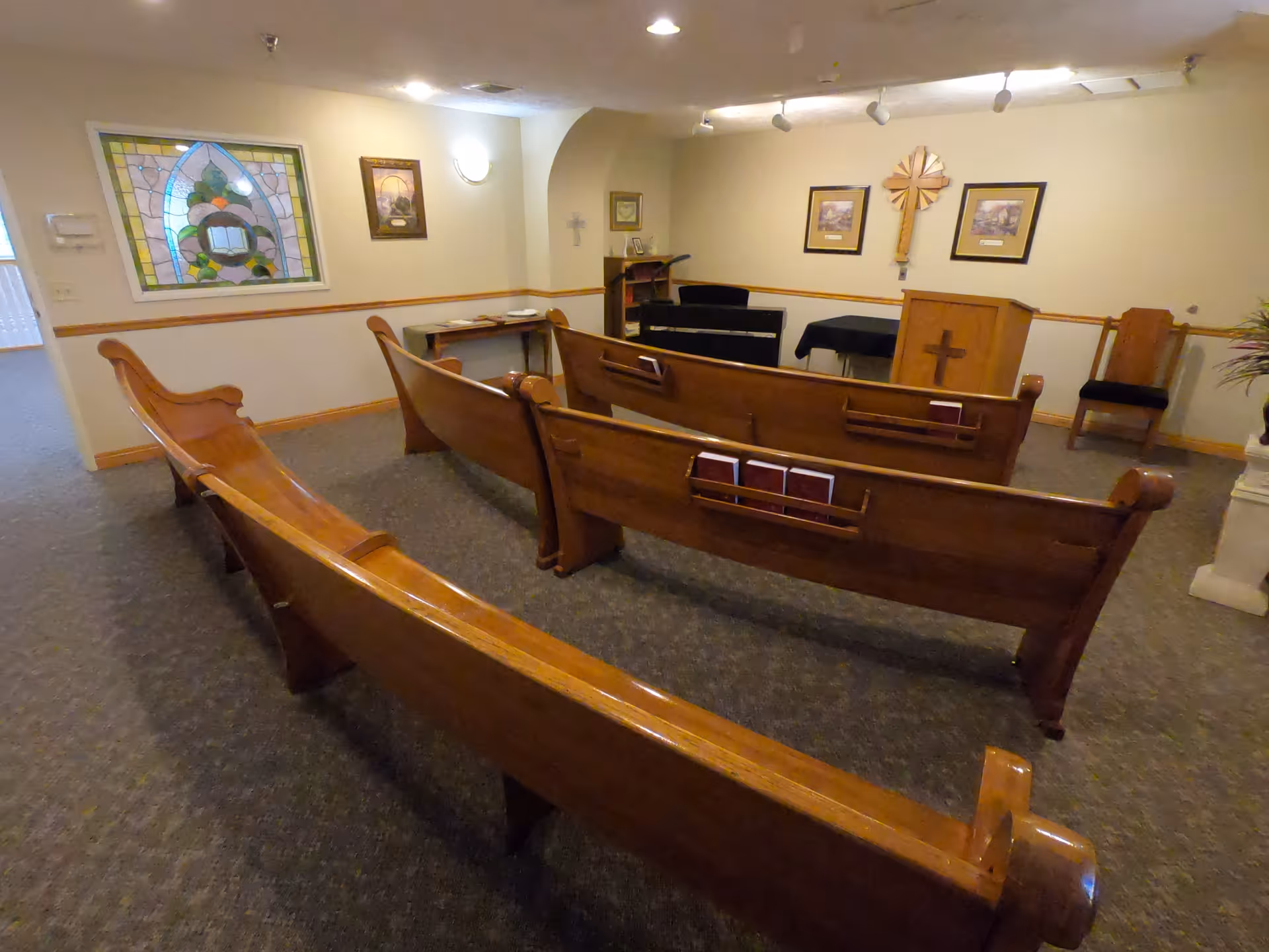 Interior view of a small chapel or prayer room with wooden pews arranged in rows facing a podium and a cross on the wall. The room has carpeted floors, framed pictures, a stained glass window, and soft lighting.