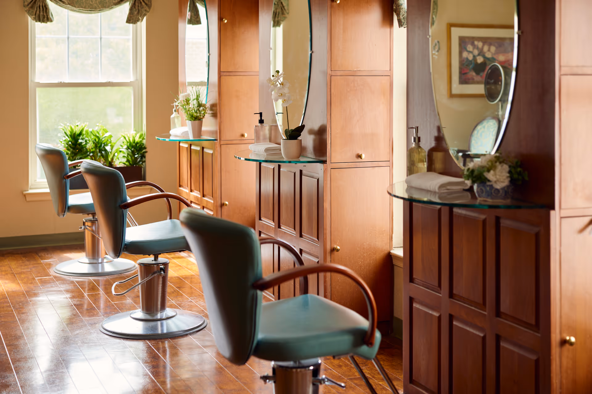 Interior view of a salon area with three teal salon chairs in front of wooden cabinets with round mirrors. The room has large windows with plants on the windowsills, and the floor is wooden.