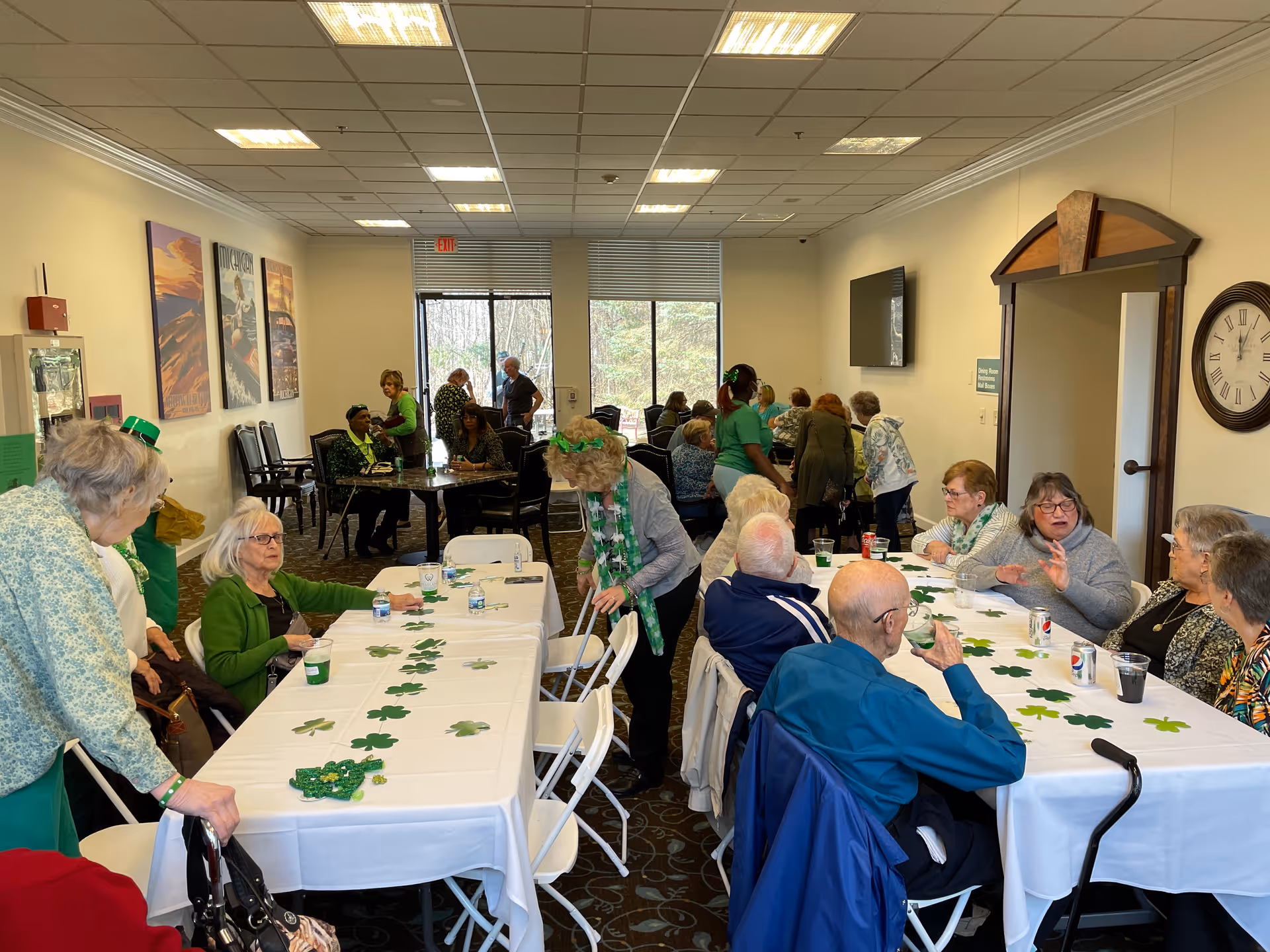 A group of elderly people gathered in a well-lit room with tables covered in white tablecloths decorated with green shamrocks. Some people are sitting and chatting while others are standing. The room has large windows with a view of trees outside, framed artwork on the walls, and a clock near a doorway. The atmosphere appears social and festive.