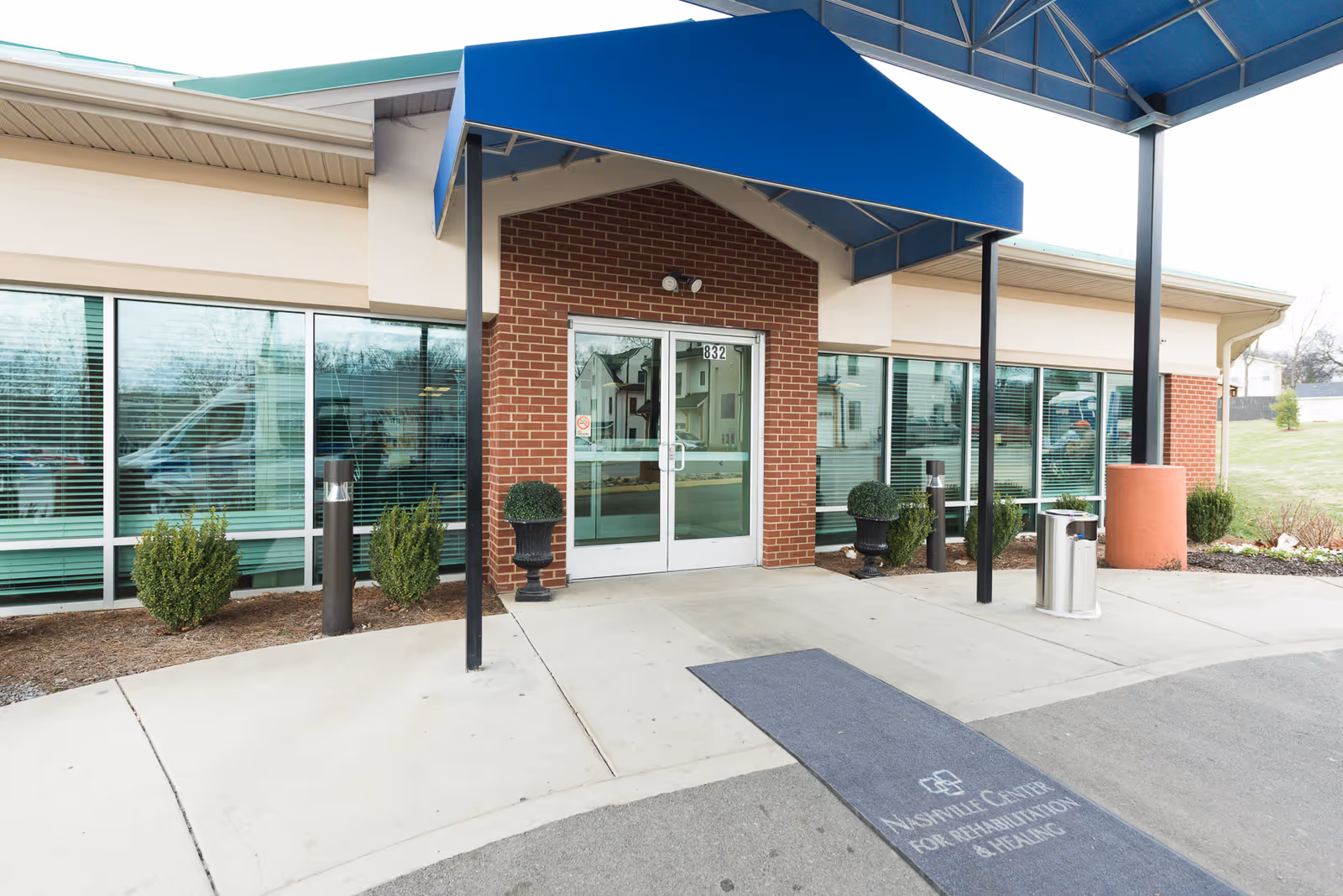 Entrance of Nashville Center for Rehabilitation & Healing featuring double glass doors under a blue awning, with brick walls and large windows on either side. There are small shrubs and decorative plants near the entrance and a mat on the ground with the facility's name.