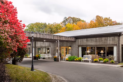Exterior view of Elderwood Village at Vestal showing a single-story building with large windows and a covered entrance. White benches and potted plants are placed near the entrance. Trees with autumn foliage surround the building.