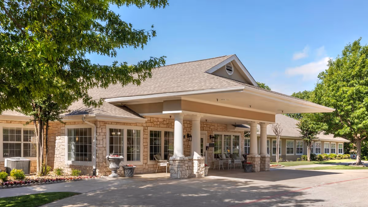 Exterior view of a single-story senior living facility building with stone and beige siding, large windows, and a covered entrance supported by white columns. There are trees and landscaping around the building under a clear blue sky.