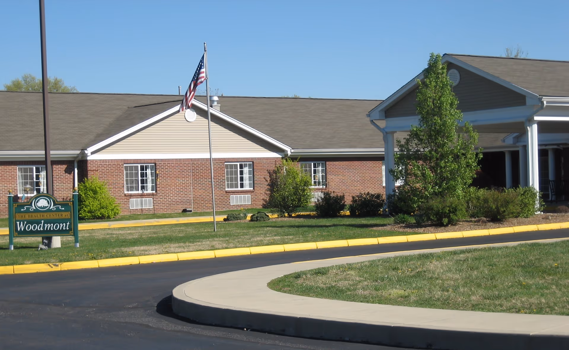 Front entrance of a single-story brick senior living building with a flagpole and a 'Woodmont' sign on the lawn.