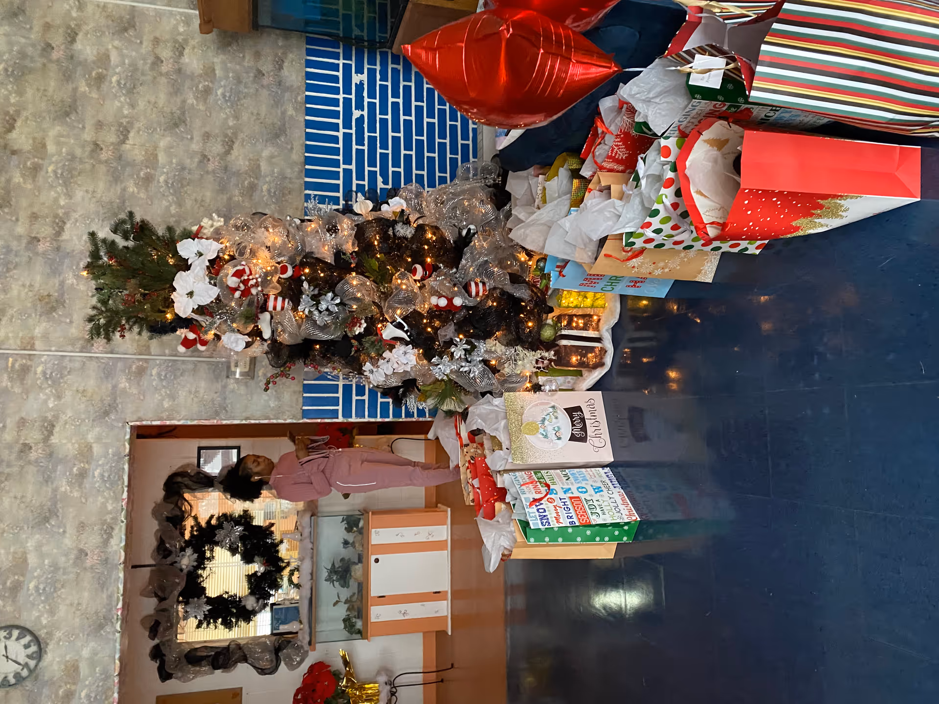 A decorated Christmas tree with white and red ornaments and lights stands in a room with a blue floor and a blue and white brick wall. Numerous wrapped Christmas presents are arranged around the base of the tree. A woman in pink clothing is standing near a doorway in the background, and a black wreath is hanging on the wall behind her.