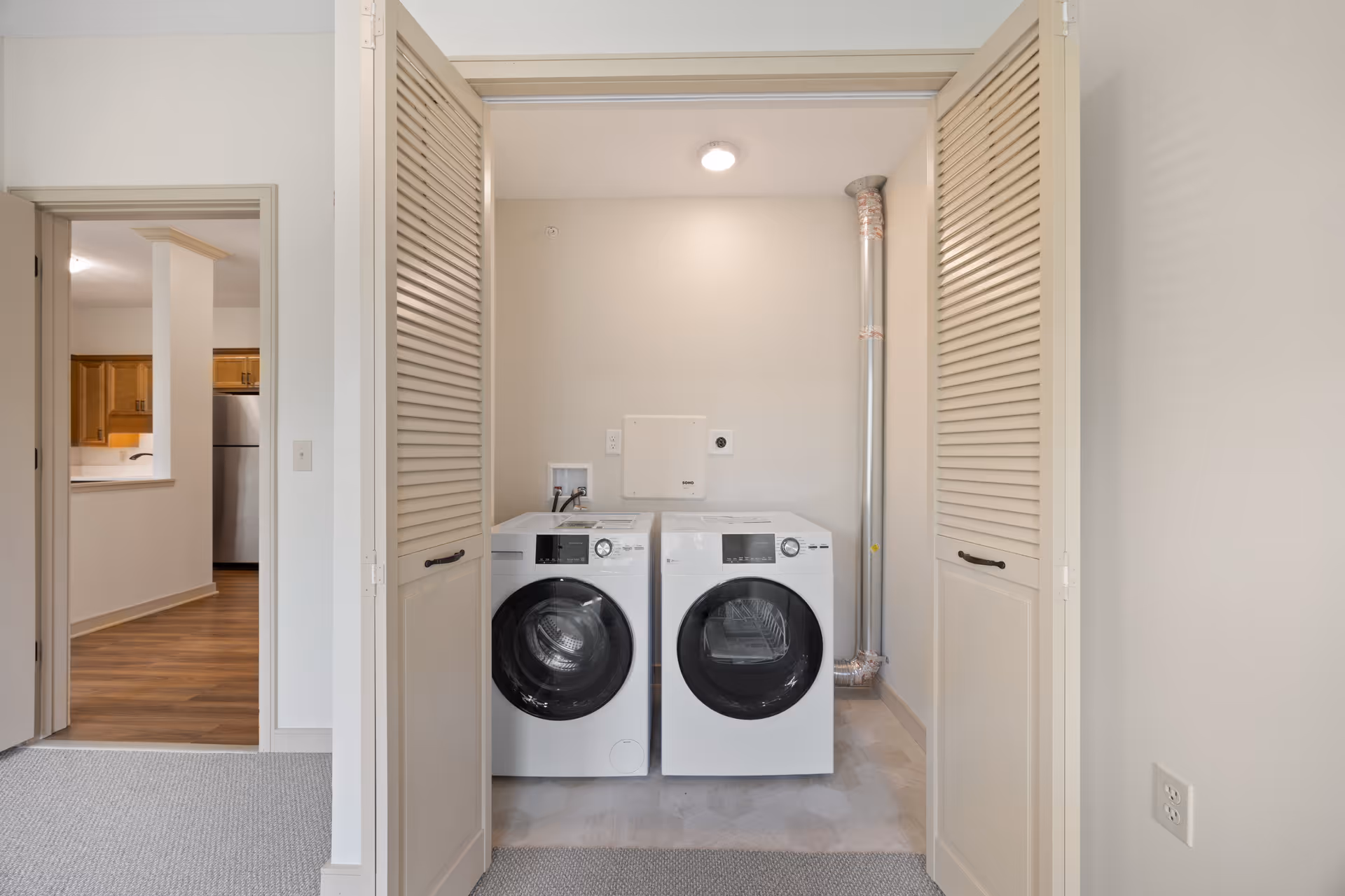 Laundry area with a front-loading washing machine and dryer side by side, enclosed by beige louvered doors. The area is well-lit with a ceiling light and has a vent pipe on the right wall. Adjacent room with wooden flooring and kitchen cabinets is visible through an open doorway on the left.