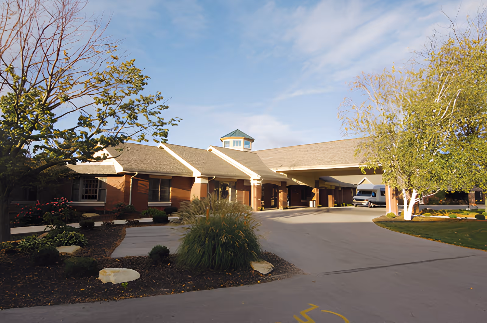 Exterior view of Good Shepherd Home facility showing a single-story building with a covered entrance driveway, surrounded by landscaped greenery including trees, bushes, and flowers under a blue sky with some clouds.