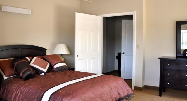 A bedroom with a bed featuring a dark wooden headboard and maroon bedding with decorative pillows. There is a white door partially open leading to another room, a lamp on a bedside table, and a dark wooden dresser with a mirror on the right side.