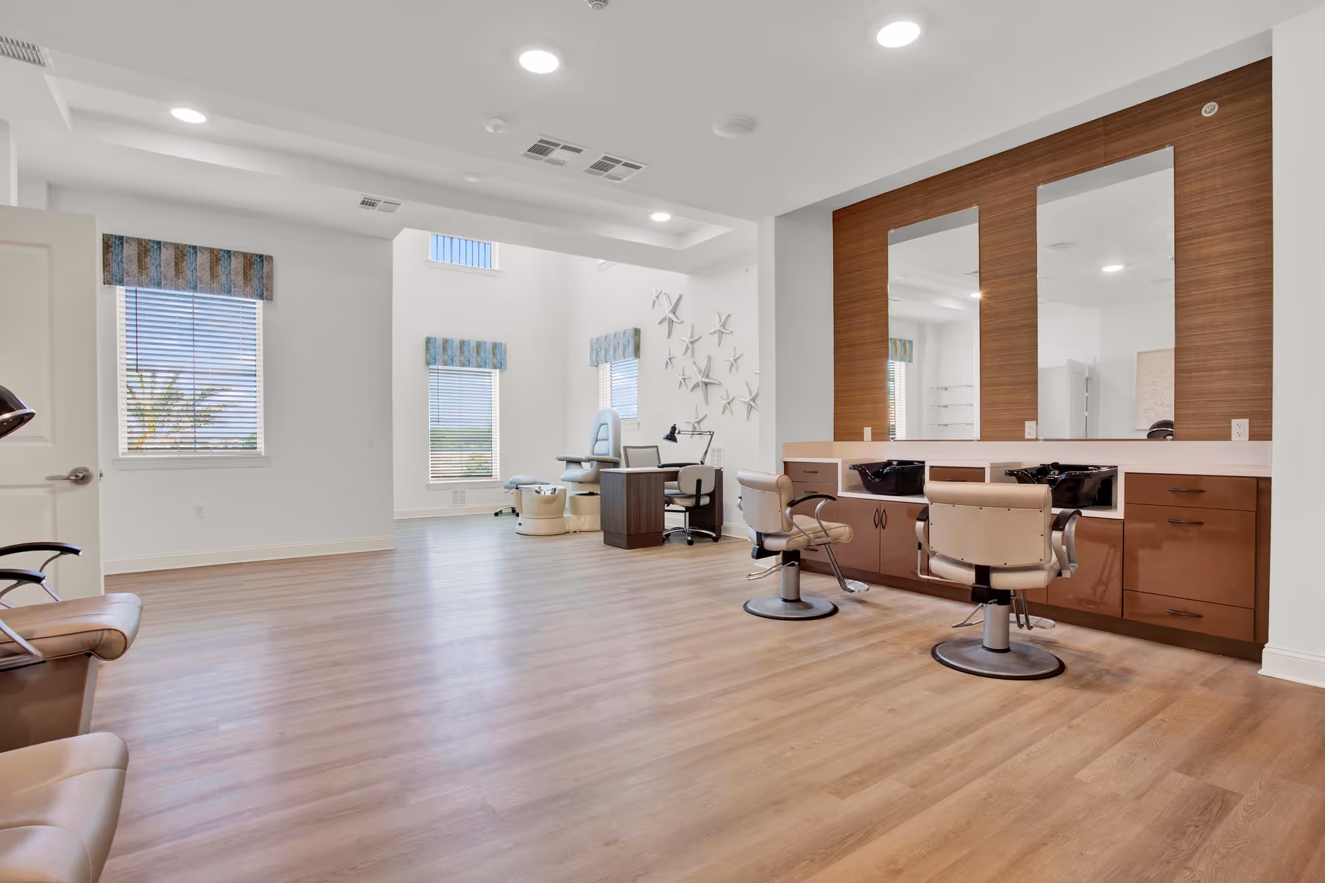 A spacious and well-lit salon area in a senior living facility with two salon chairs in front of large mirrors mounted on a wooden panel. The room features light wood flooring, white walls, and several windows with striped valances allowing natural light to enter. In the background, there is a pedicure chair and a small desk with a chair. Decorative starfish adorn one wall.