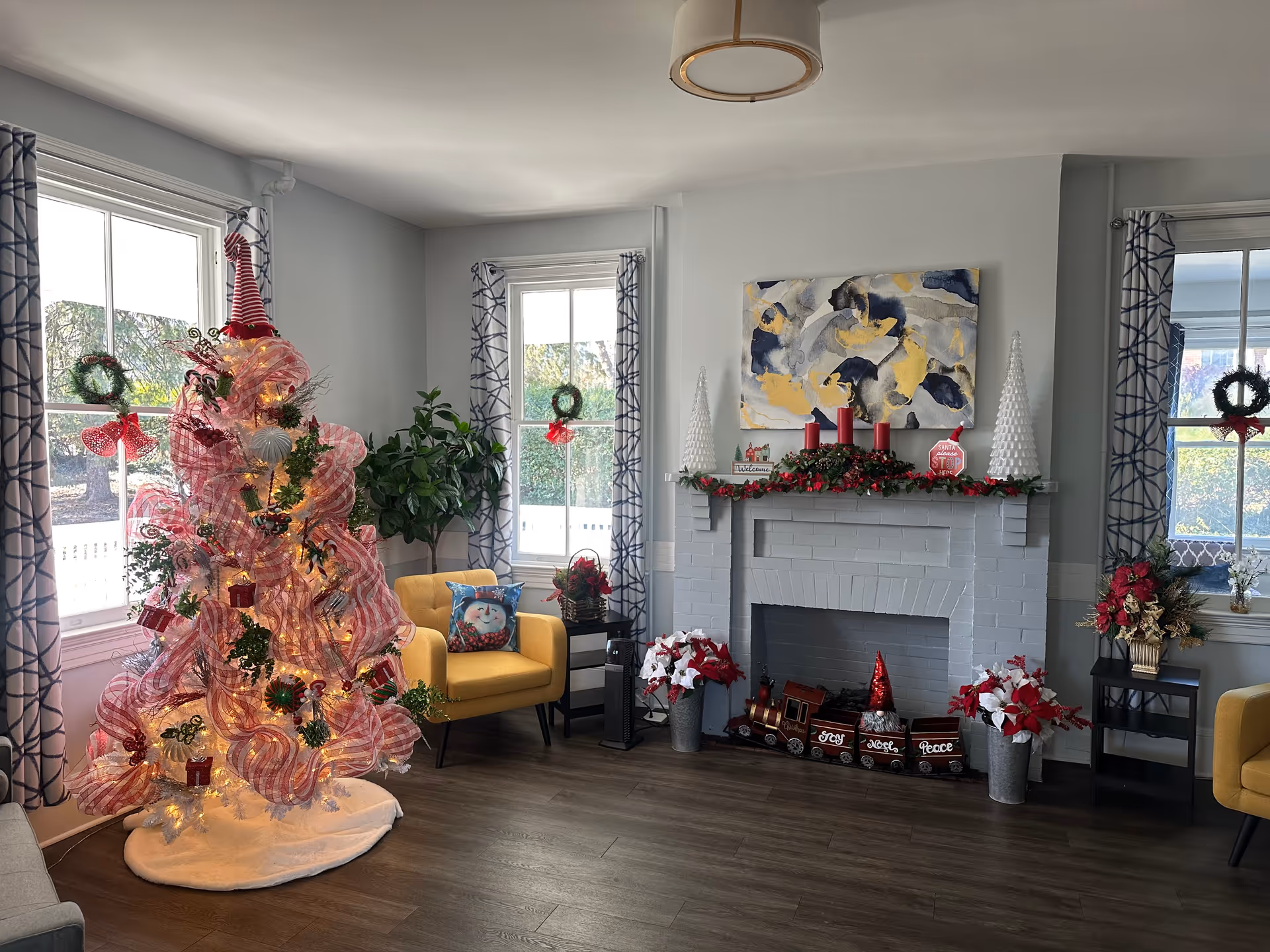 A cozy living room decorated for Christmas with a white Christmas tree wrapped in pink ribbon and adorned with ornaments, a yellow armchair with a snowman pillow, a white brick fireplace decorated with red candles, garlands, and holiday figurines, and windows with patterned curtains and small wreaths.