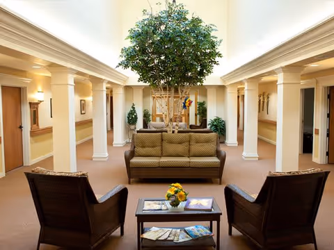 A bright and spacious common area with a large indoor tree in the center. The room features a brown cushioned sofa and two wicker armchairs arranged around a small coffee table with magazines and a flower vase. The area is framed by white columns and has beige walls and carpeted floors, with doorways and framed artwork visible in the background.