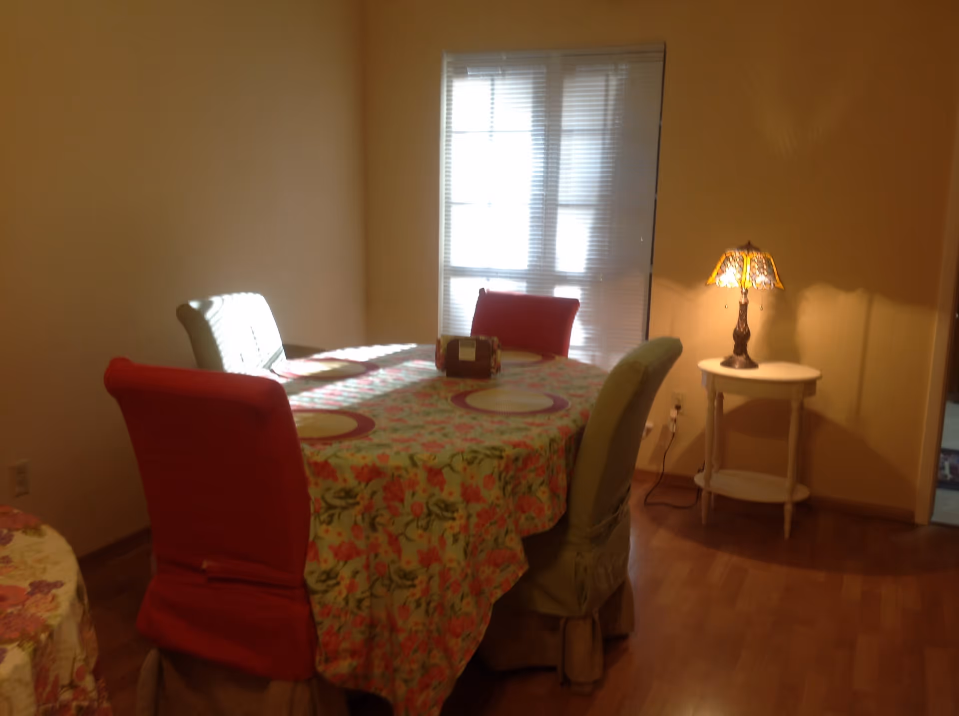 A dining room with a rectangular table covered with a floral tablecloth and four chairs with slipcovers in red, white, and green. There are placemats on the table and a small lamp on a round side table near a window with blinds.