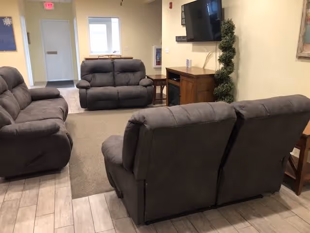 A well-lit common room with several gray reclining sofas arranged around a wall-mounted TV and a small media cabinet.
