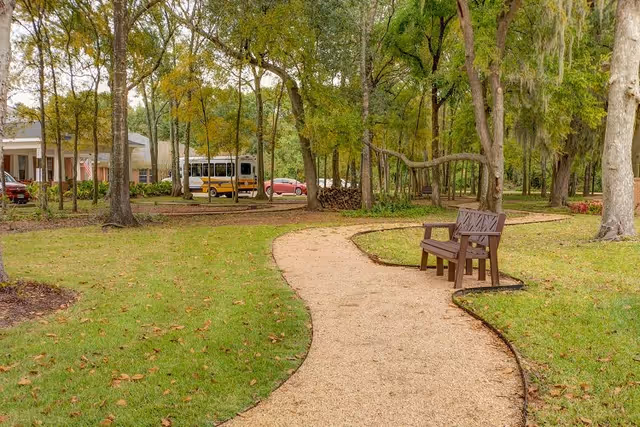 A winding gravel path through a green park-like area with tall trees and a wooden bench on the right side. In the background, there is a building partially visible with a school bus and a red car parked nearby.