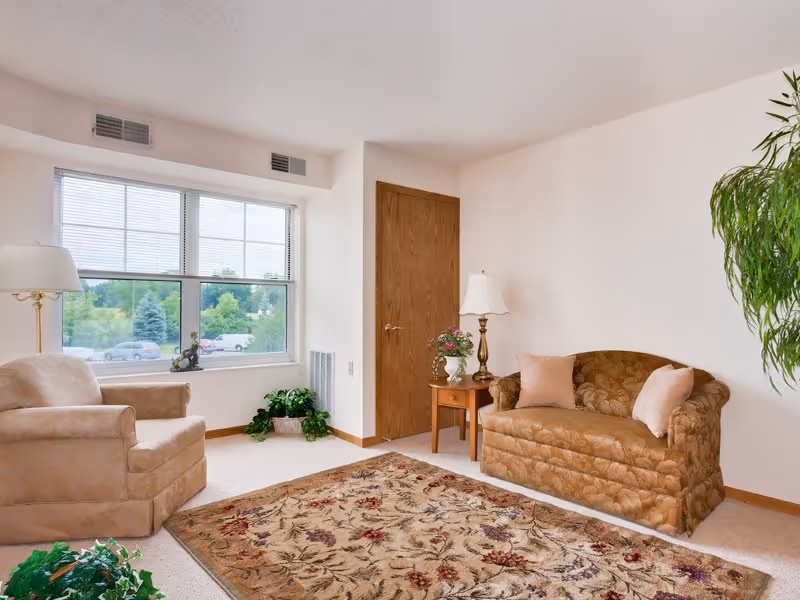 A cozy living room with a large window showing greenery outside. The room features a beige armchair, a brown patterned loveseat with two light pink pillows, a wooden side table with a lamp and a flower vase, a floral area rug, and a tall green plant in the corner.