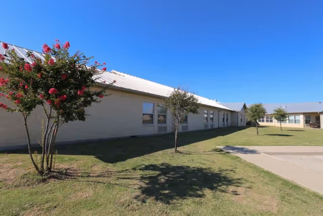 Exterior view of a single-story building with light-colored walls and a metal roof, surrounded by a grassy area with small trees and a clear blue sky overhead.