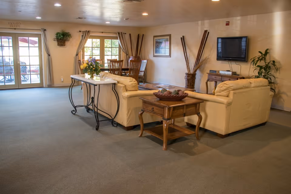 A cozy living room area in Casa De Flores featuring two beige leather sofas arranged around a wooden coffee table with decorative items. A flat-screen TV is mounted on the wall above a small wooden console table. In the background, there is a dining table with chairs near a window with curtains, and glass doors leading to an outdoor patio with seating. The room is warmly lit with recessed ceiling lights and decorated with plants and wall art.