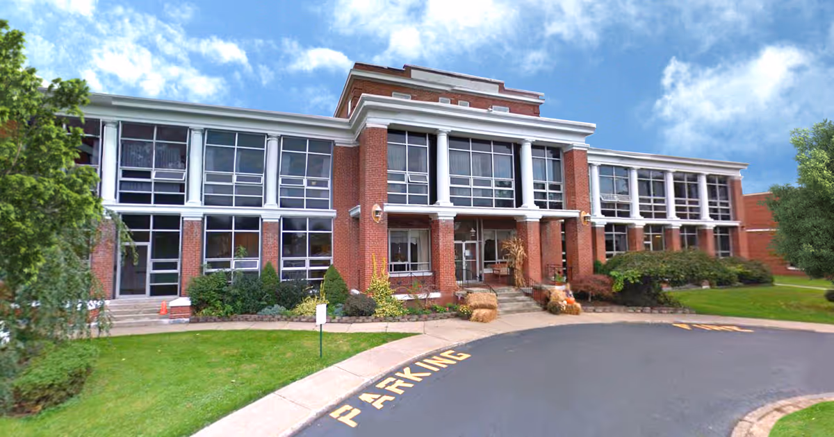 Front exterior view of Waterview Heights Rehabilitation and Nursing Center, a two-story brick building with large windows, white columns, and a curved driveway with parking markings. The building is surrounded by green grass, shrubs, and trees under a partly cloudy sky.