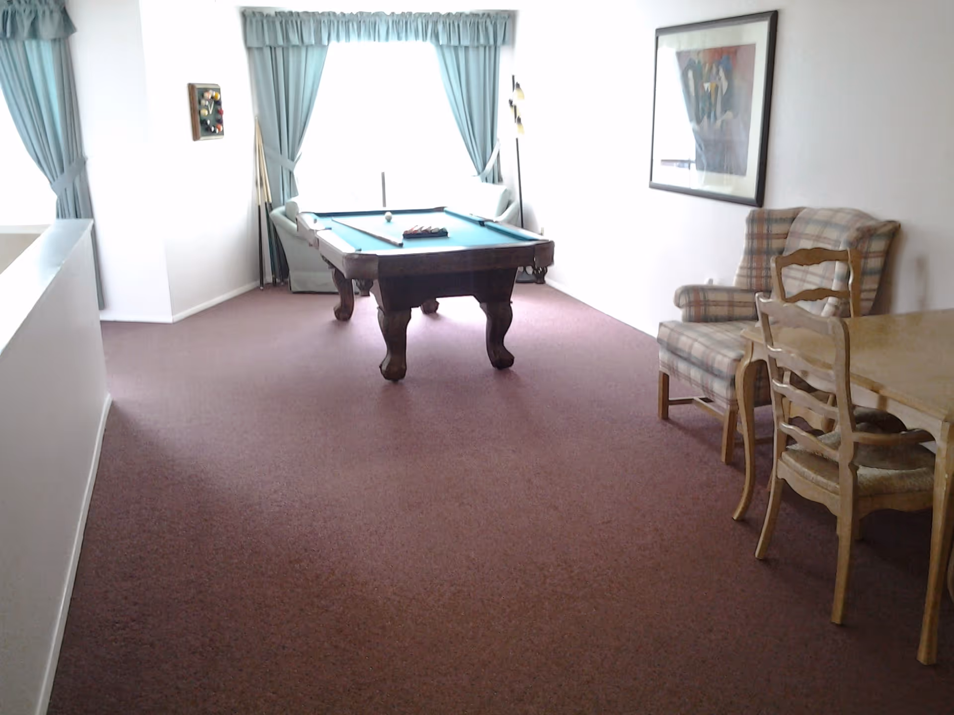A room with a pool table in the center, two armchairs near a large window with blue curtains, a framed picture on the wall, and a wooden dining table with chairs on the right side.