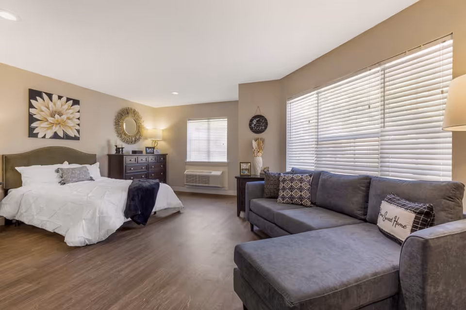 Bright, spacious studio-style bedroom with a bed, dresser, and a gray sectional sofa next to large blinds-covered windows.