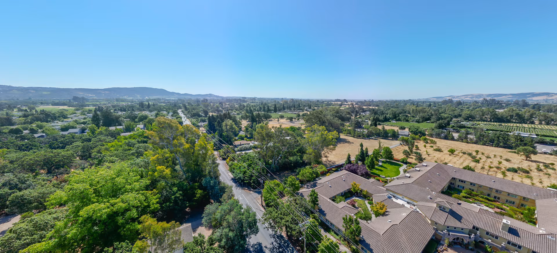 Aerial view of a senior living facility named Cogir of Sonoma Plaza surrounded by trees, roads, and open fields under a clear blue sky.