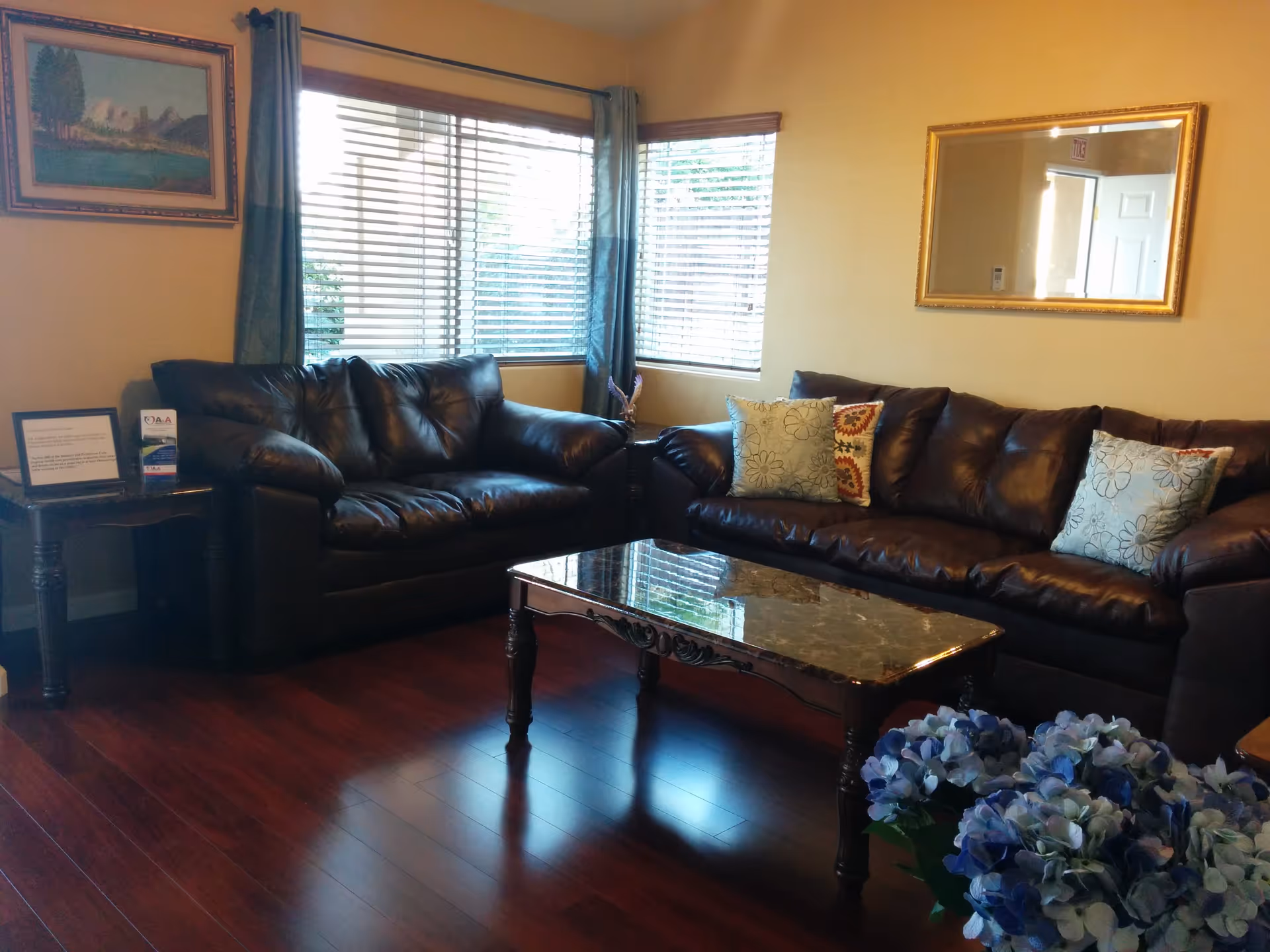 A cozy living room with two dark brown leather sofas, a marble-top coffee table, and a side table with framed documents and brochures. The room has wooden flooring, a large window with blinds and blue curtains, a framed landscape painting on the wall, a large mirror, and a bouquet of blue and white flowers in the foreground.