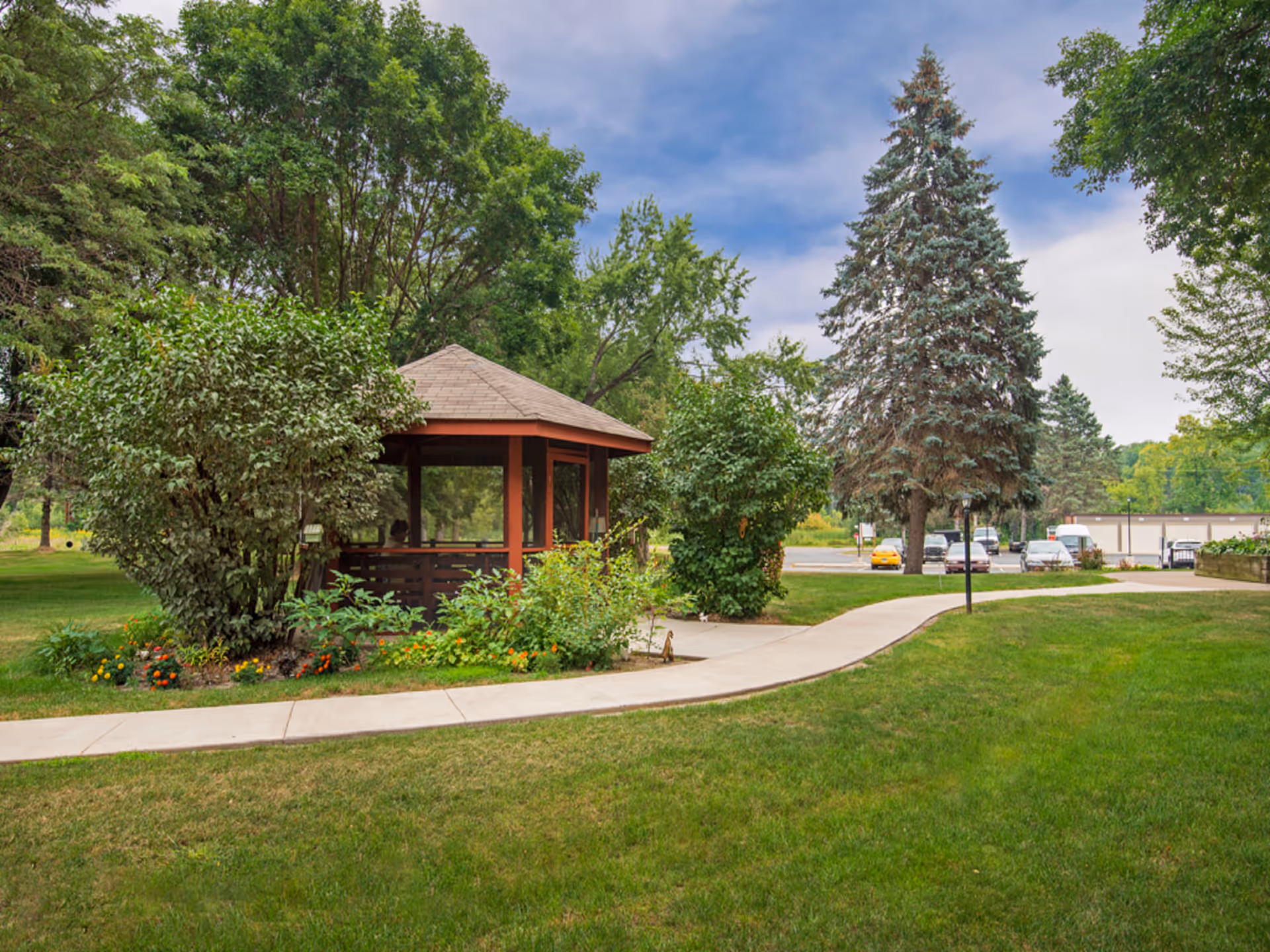 A peaceful outdoor area at Kilkenny Court featuring a small gazebo surrounded by lush green trees and bushes, a curved concrete pathway, and a grassy lawn. Several cars are parked in the background near a building under a partly cloudy sky.