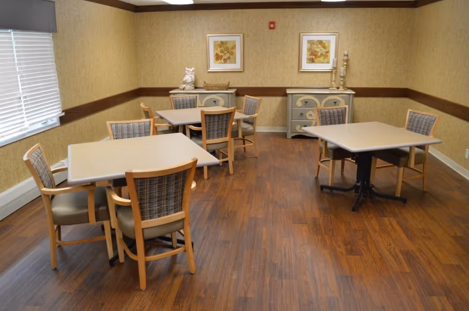 A small dining room with three square tables, each surrounded by four wooden chairs with cushioned seats and backs. The room has wood flooring, beige patterned wallpaper, and two framed floral artworks on the far wall above two decorative dressers. A window with closed blinds is on the left side.