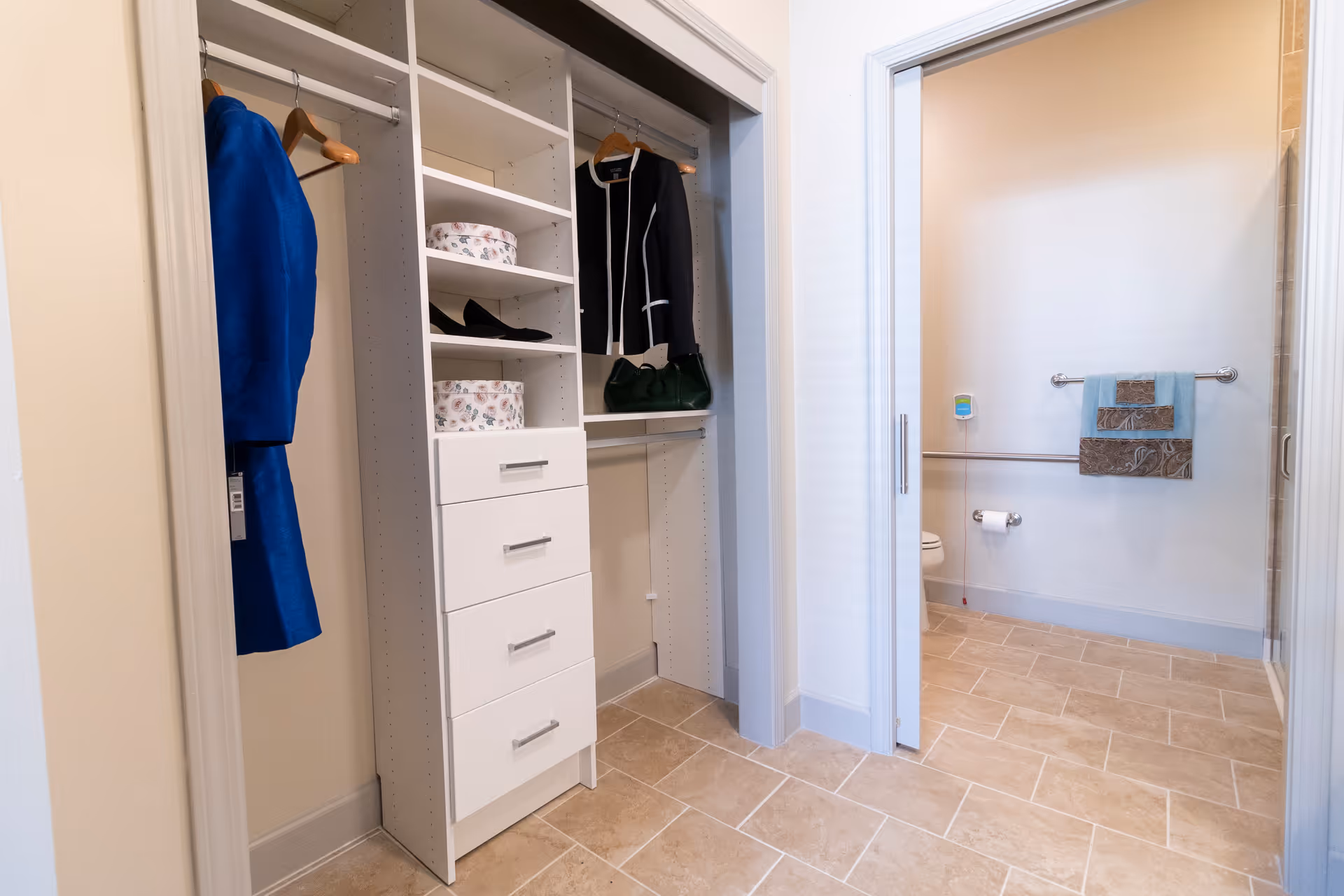 A white built-in closet with shelves and hanging garments opens to a tiled, accessible bathroom with grab bars and a towel on the wall.