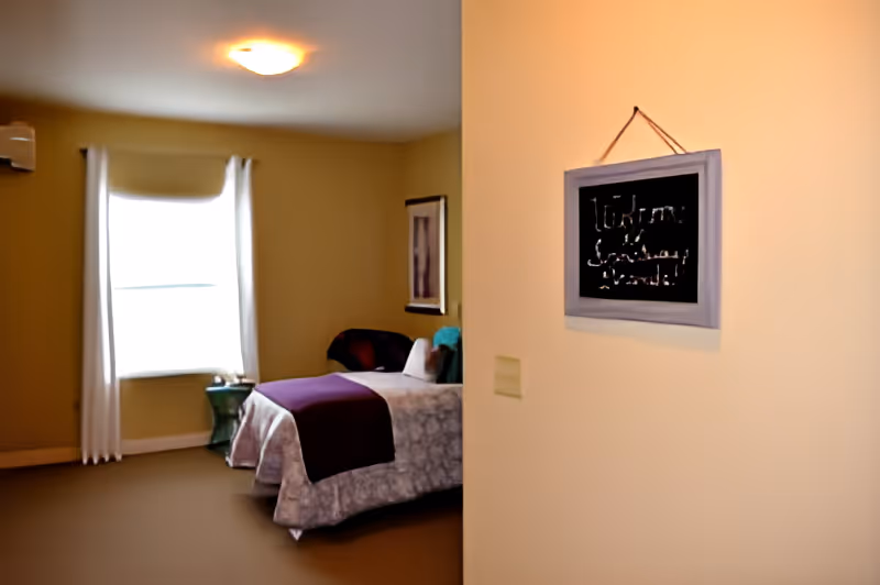 A cozy bedroom with a single bed covered in a light-colored bedspread and a dark purple throw blanket. There is a window with white curtains letting in natural light, a small green side table next to the bed, and a framed picture on the wall. A small chalkboard sign hangs on the wall near the entrance with the words 'Welcome Sanctuary Grande!' written on it.