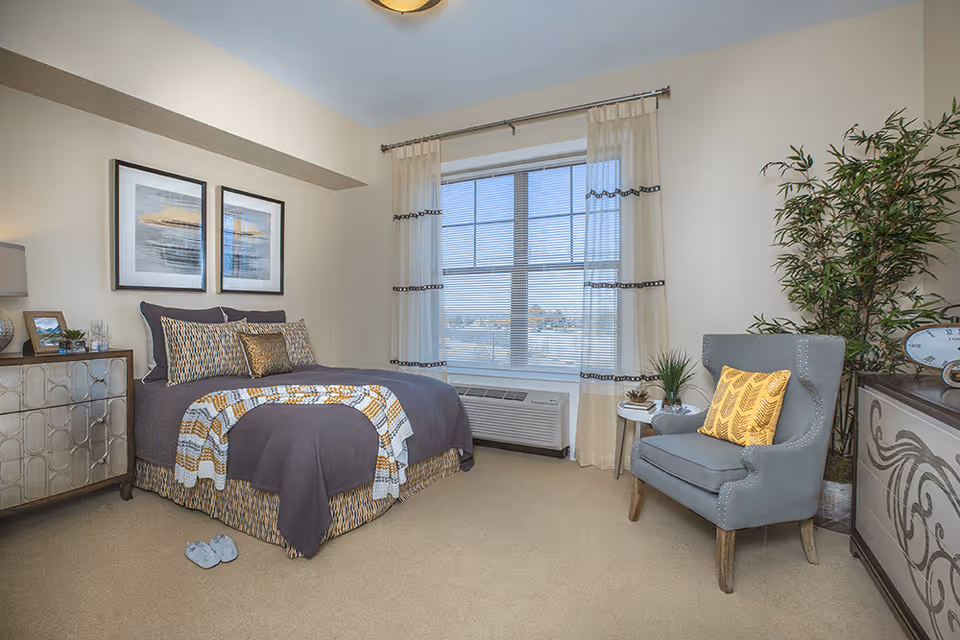 A cozy bedroom with a neatly made bed featuring a gray and yellow patterned comforter and pillows. There are two framed pictures hanging above the bed. To the left of the bed is a decorative dresser with a lamp and small decor items. A large window with sheer curtains lets in natural light. On the right side, there is a gray armchair with a yellow pillow, a small round side table with plants, and a large potted plant in the corner. The room has beige walls and carpeted floor.