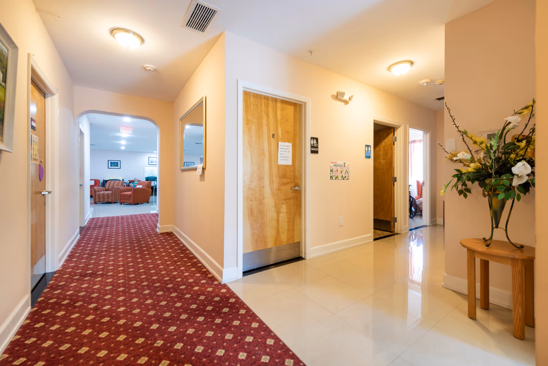 Interior hallway of Sunny Days Assisted Living Facility with a red patterned carpet leading to a common area with chairs. The hallway has beige walls, wooden doors, a mirror on the wall, and a small table with a vase of flowers on the right side.