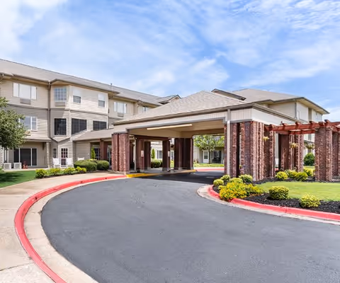 Covered porte-cochere and circular driveway at the front of a multi-story residential building with landscaped beds under a blue sky.