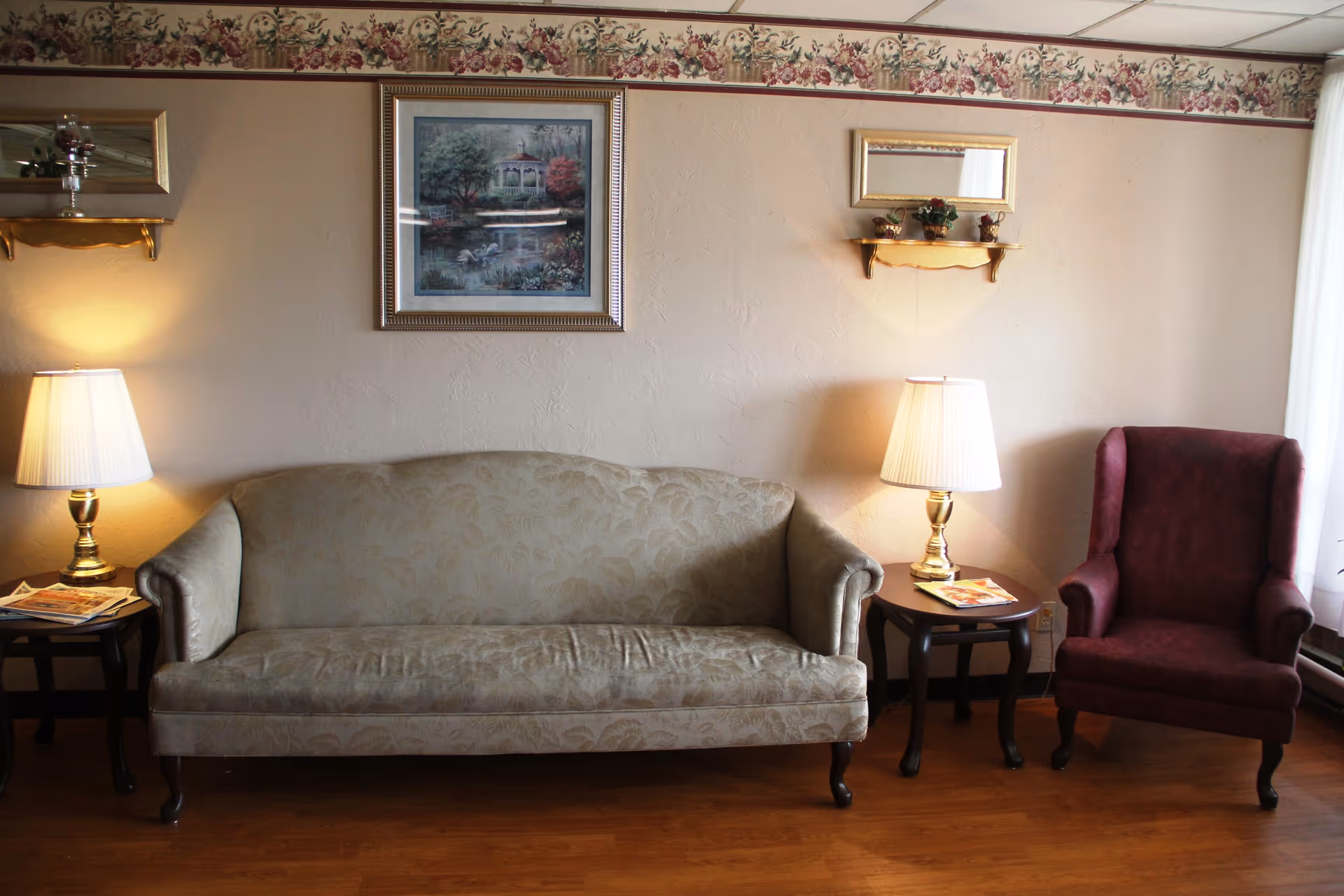 A cozy living room area with a beige patterned sofa centered between two wooden side tables, each holding a brass lamp with a white lampshade. To the right of the sofa is a burgundy upholstered armchair. Above the sofa hangs a framed painting of a gazebo by a pond, and above the right side table is a small wall shelf with decorative items and a small mirror. The room has light-colored walls with a floral wallpaper border near the ceiling and wooden flooring.