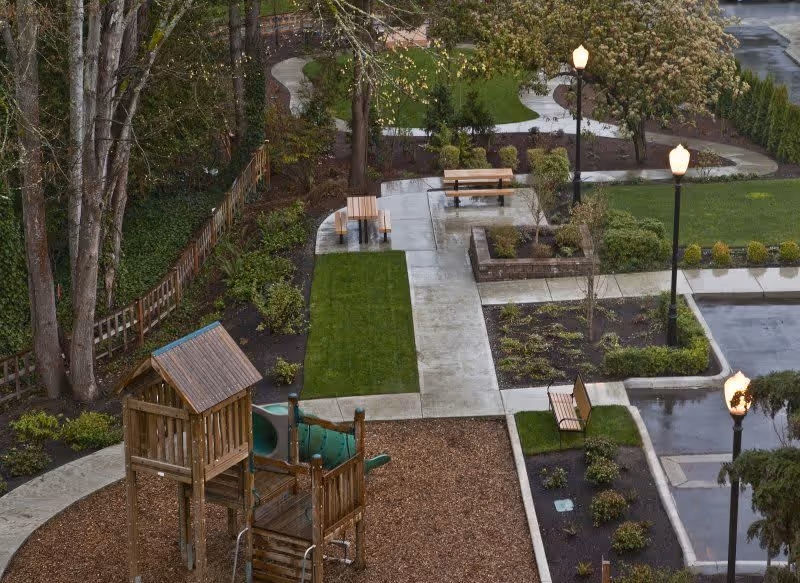 Outdoor area with a wooden playground structure, benches, paved walkways, green grass patches, trees, and street lamps. The scene appears to be part of a landscaped garden or park area within a facility.