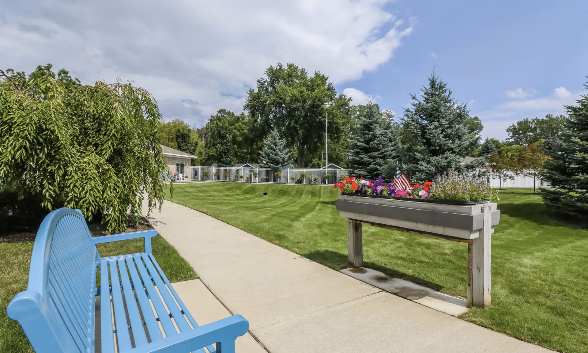 Outdoor garden area with a blue bench on the left side of a concrete pathway, green grass, various trees, and a raised flower bed with colorful flowers and a small American flag under a partly cloudy sky.