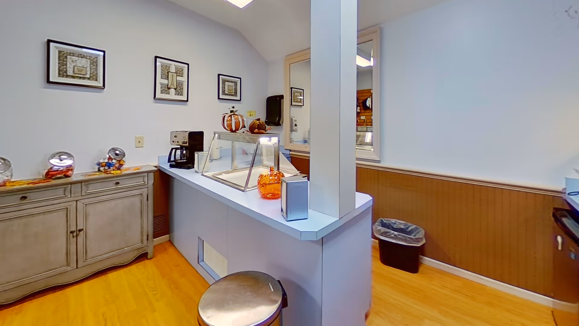 Interior view of a small serving area with a counter, a coffee maker, decorative pumpkins, and a glass display case. There is a wooden cabinet with jars of candy on top, three framed pictures on the wall, a trash can, and a paper towel dispenser mounted on the wall.