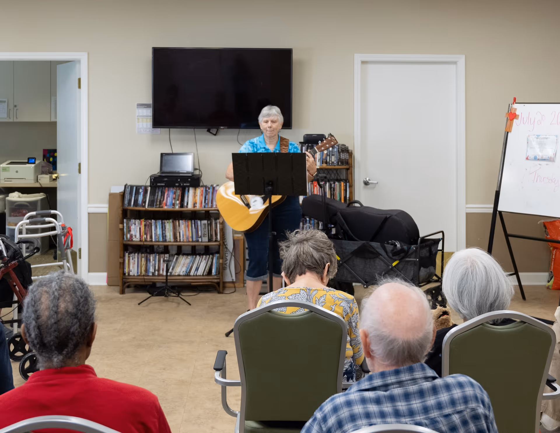 An elderly woman playing an acoustic guitar and singing in front of a seated audience in a room with bookshelves, a large TV mounted on the wall, and a whiteboard with writing on it.