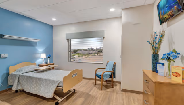 A clean and well-lit bedroom in a healthcare facility with a single hospital-style bed, a bedside table with a lamp and flowers, a wooden chair with a blue cushion, a window with blinds showing an outside view, a dresser with decorative vases and cards, and a wall-mounted TV.