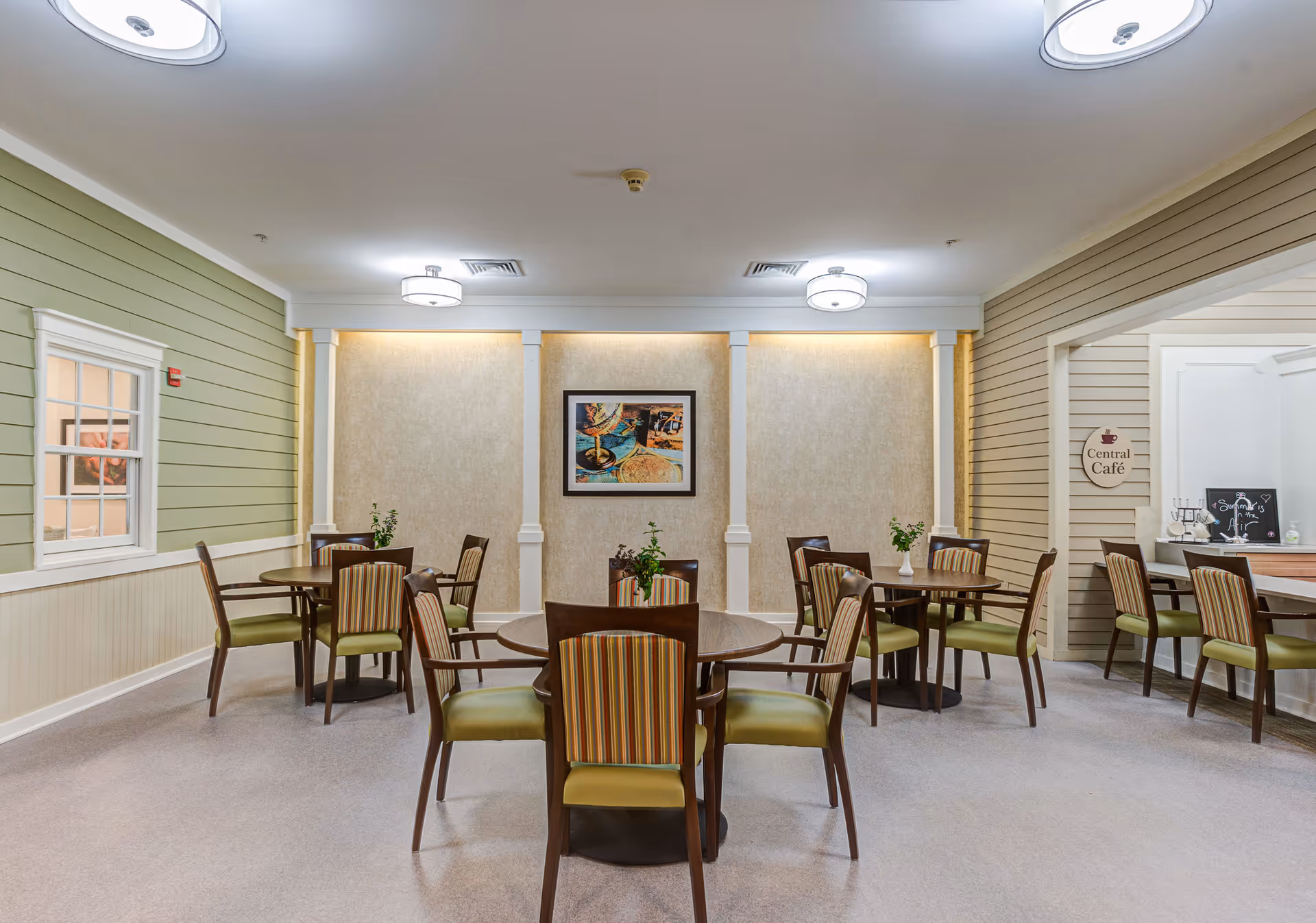 A cozy dining area with several round wooden tables and chairs featuring green cushions and striped backs. The walls are painted in soft green and beige tones with white trim. A framed picture hangs on the far wall, and there are small plants on the tables. To the right, there is a sign that reads 'Central Café' above a counter area with additional seating.
