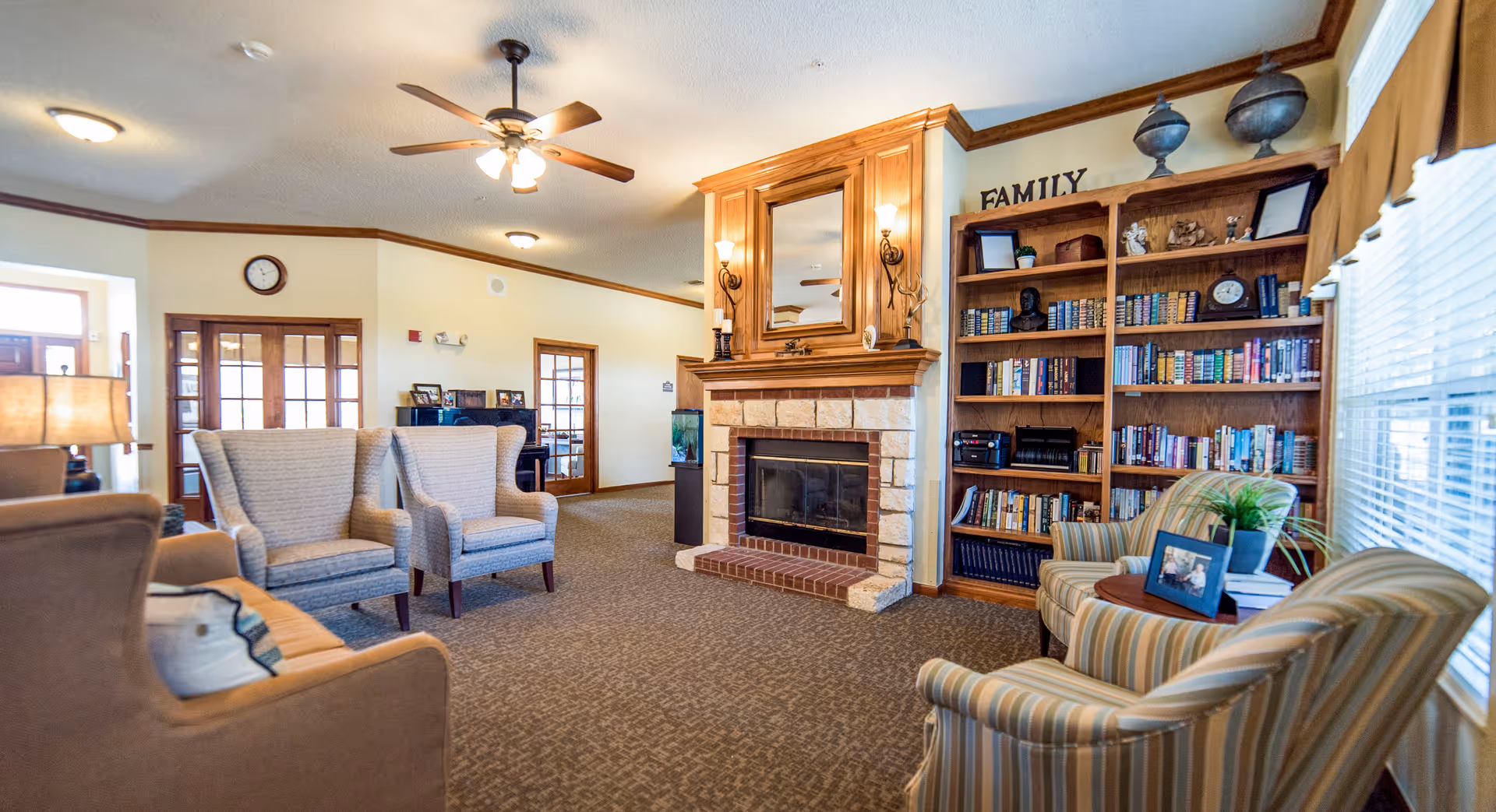 A cozy living room area in Brookdale Mansfield featuring a stone and wood fireplace with a mirror above it, surrounded by comfortable armchairs and a sofa. There is a wooden bookshelf filled with books and decorative items, a small round table with framed photos and a plant, and large windows with blinds letting in natural light. The room has beige walls, carpeted floor, and ceiling fans with lights.