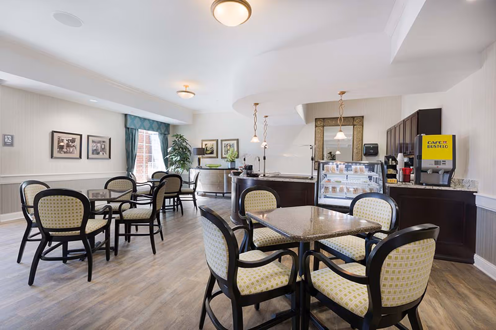 Bright community dining area with several tables and upholstered chairs, a serving counter with a display case and coffee station.