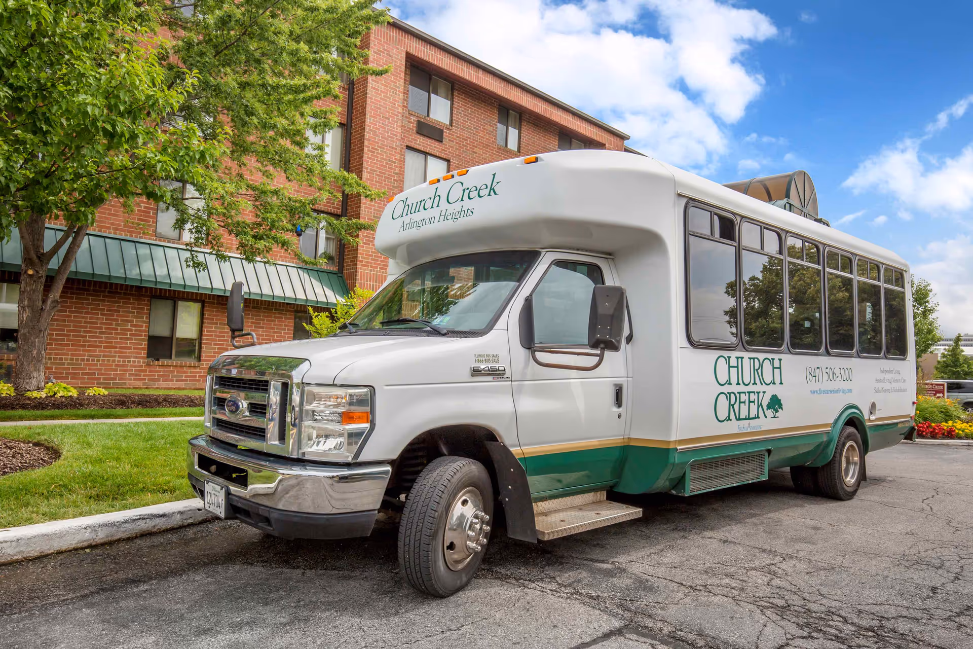 A white and green Church Creek shuttle bus parked in front of a brick building with green awnings and surrounded by trees and landscaping under a partly cloudy sky.